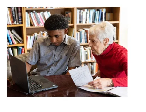 A teenage boy shows a laptop screen to an older woman. Both are sitting at a table in front of a bookshelf.